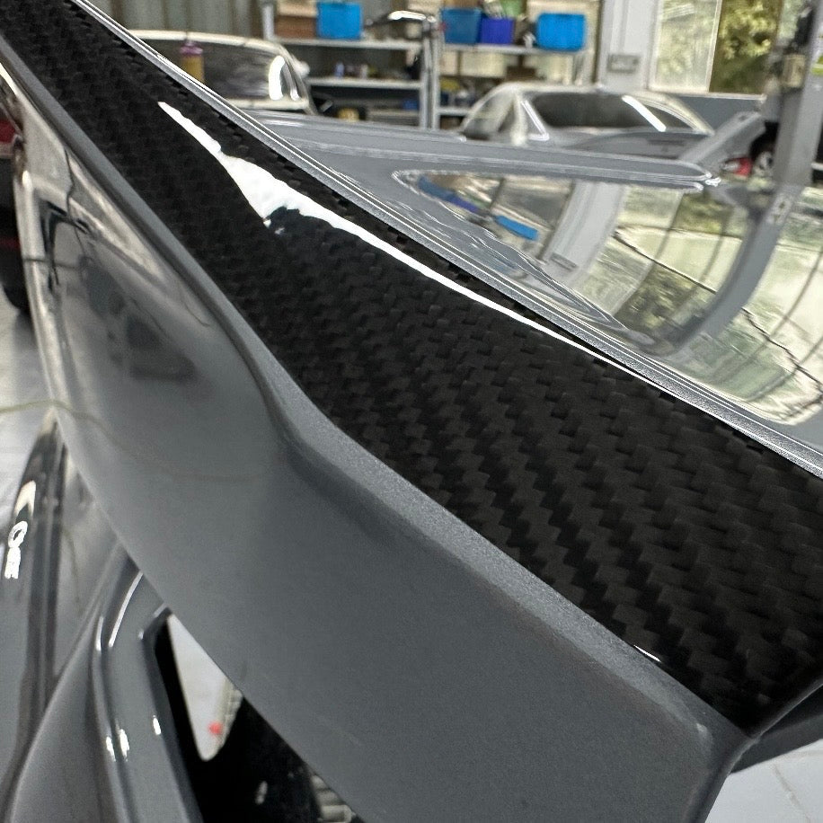 Detail of a BMW G87 M2 CS carbon fiber trunk lid, grey OEM paint meeting exposed clear coated carbon weave along the raised ducktail edge