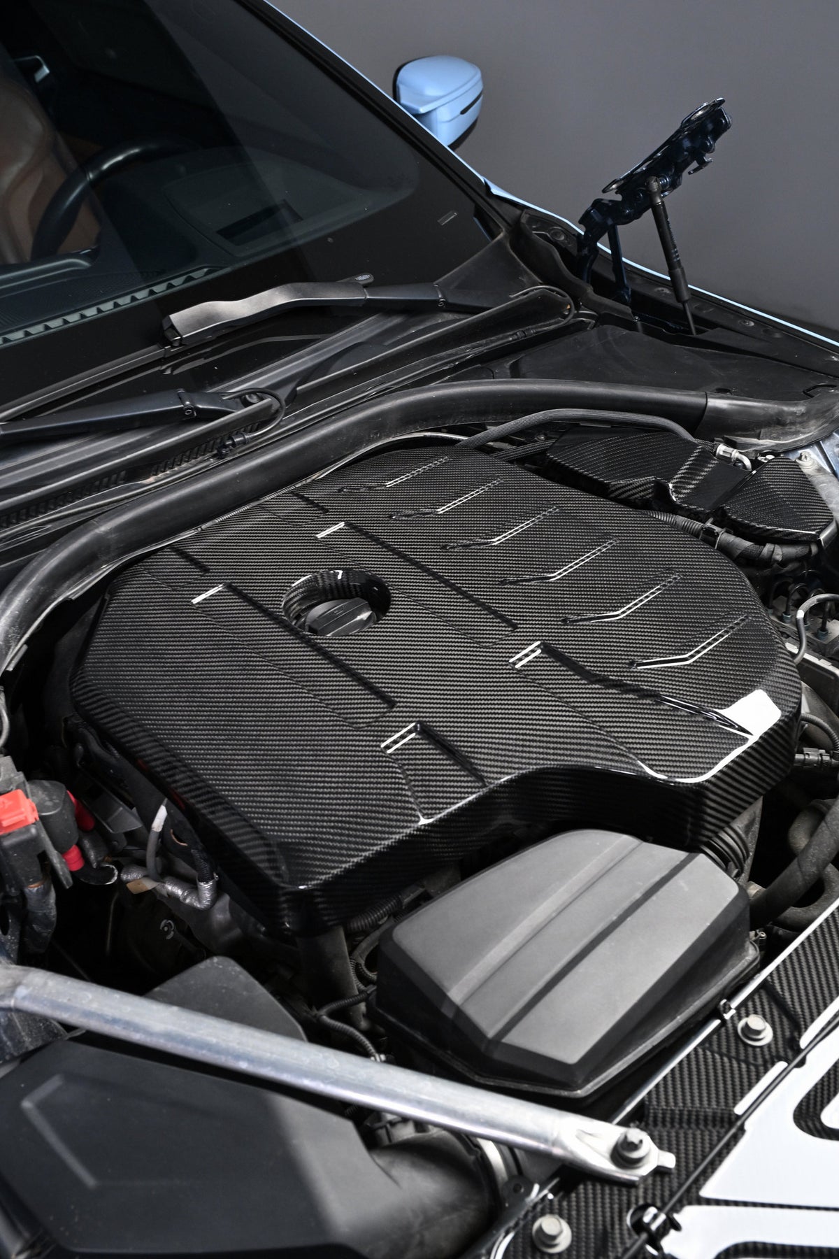 BMW G22 4 Series B48 carbon fiber engine cover is seen at an angle behind a silver metal bar in a blue car.