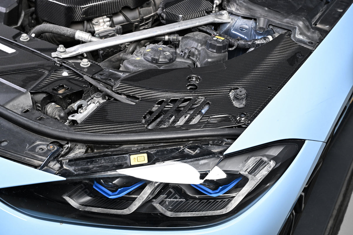 BMW G22 4 Series carbon fiber headlight cover panels are seen above a blue car's headlight in a low angled view.