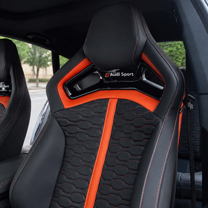 Audi RS Sport Bucket Seat backrest – black leather with orange vertical accent stripes, wave-quilted center and Audi Sport checkered badge on shoulder bridge