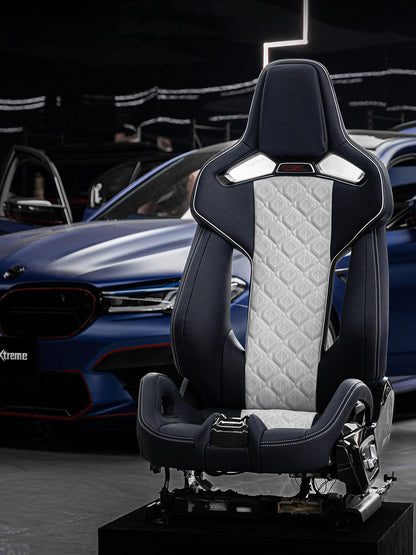 BMW M5 carbon fiber bucket seat on a display stand in navy and white diamond quilted bespoke leather, illuminated M headrest emblem and exposed carbon fiber shell, blue BMW M5 in the showroom background