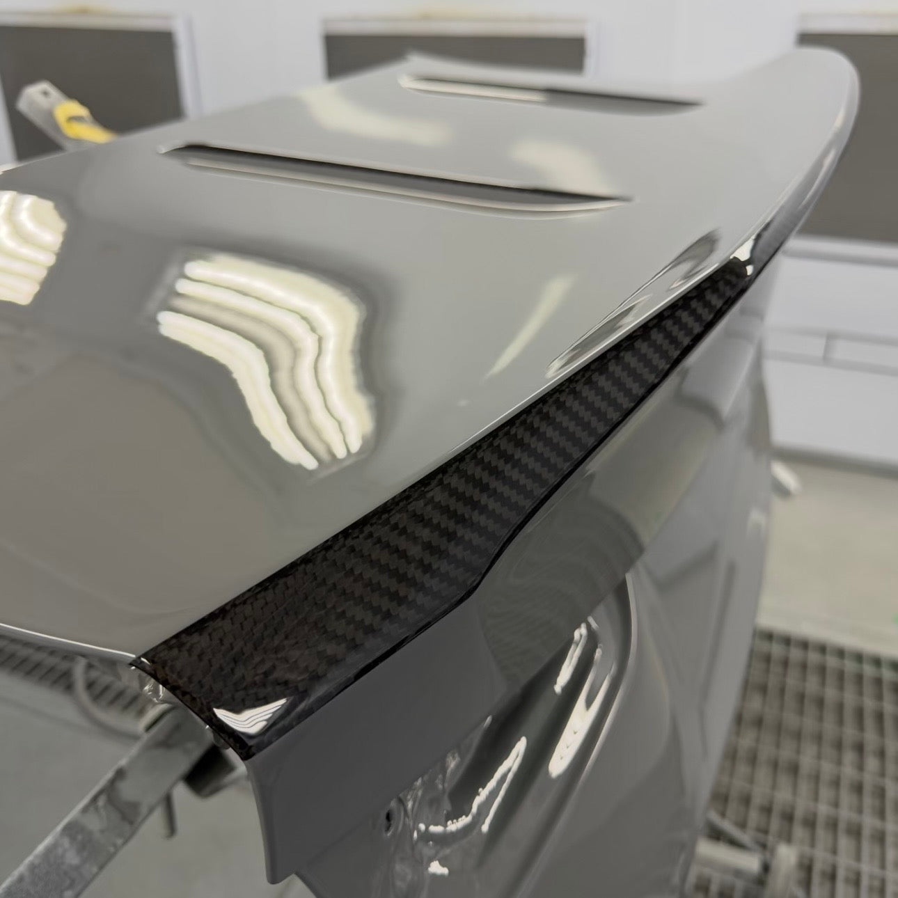BMW G87 M2 CS carbon fiber trunk lid in grey OEM paint with the ducktail spoiler lip left in exposed clear coated carbon weave, a carbon stripe finish