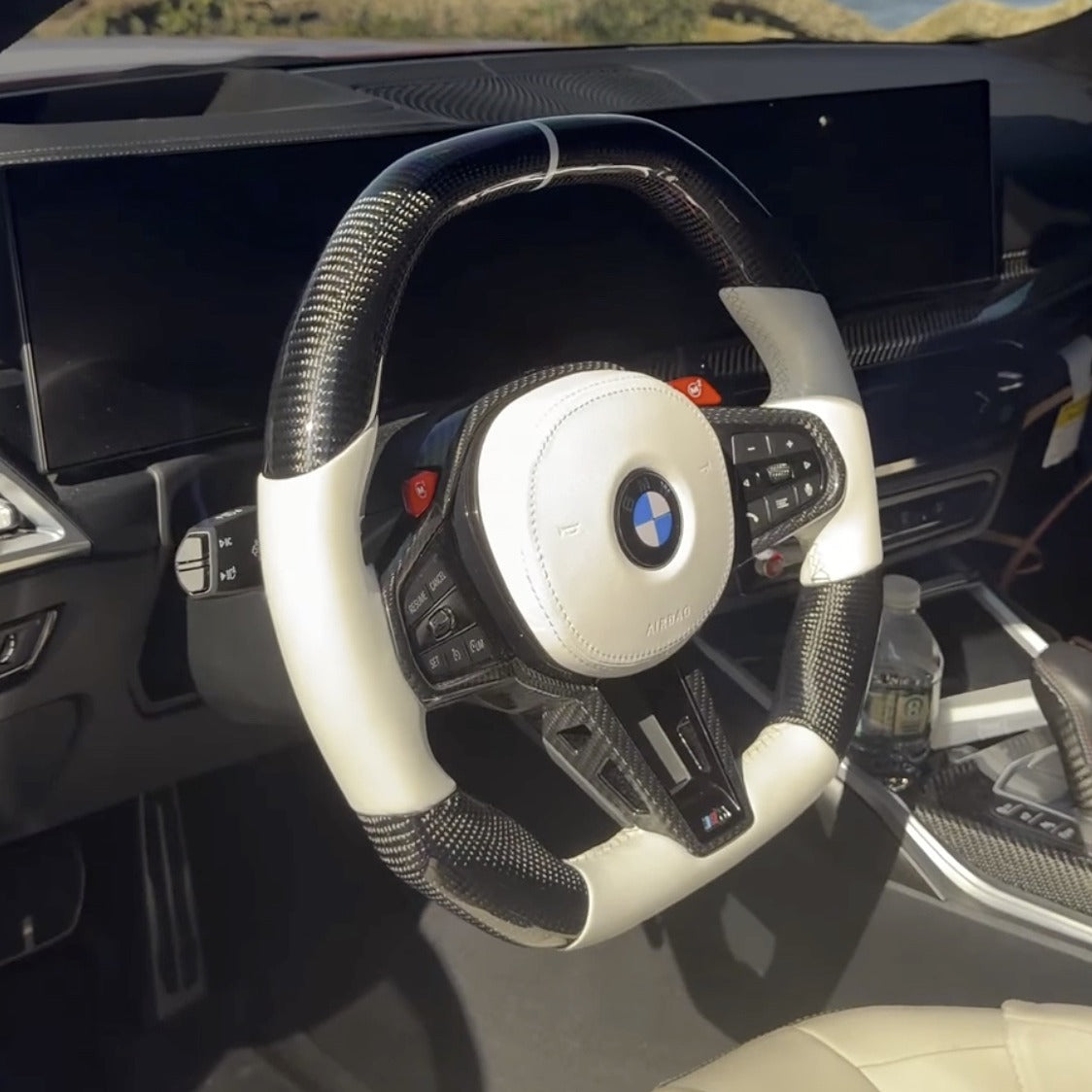 Custom carbon fiber steering wheel installed in a BMW G80 M3, white leather grip with carbon fiber center trim and red M1 M2 buttons