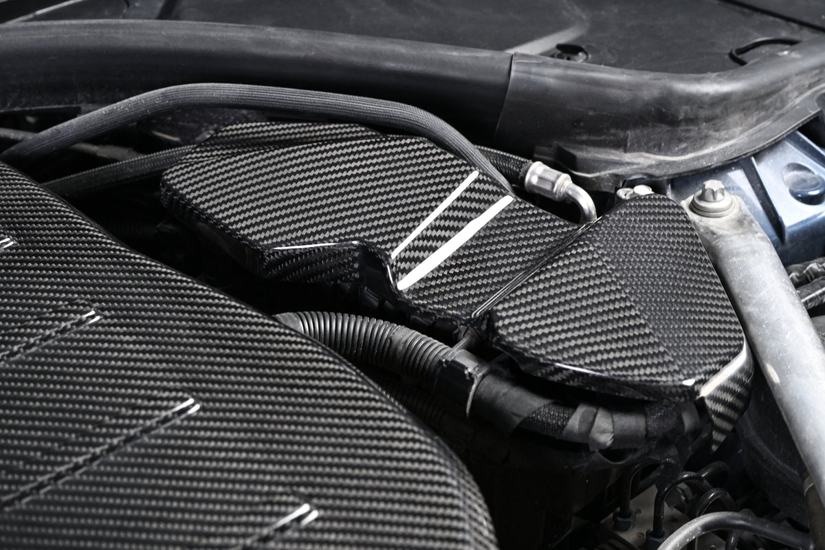 BMW G22 4 Series carbon fiber ECU battery terminal cover is seen close up beneath a curved hose in the engine bay.