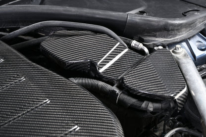 BMW G22 4 Series carbon fiber ECU battery terminal cover is seen close up beneath a curved hose in the engine bay.