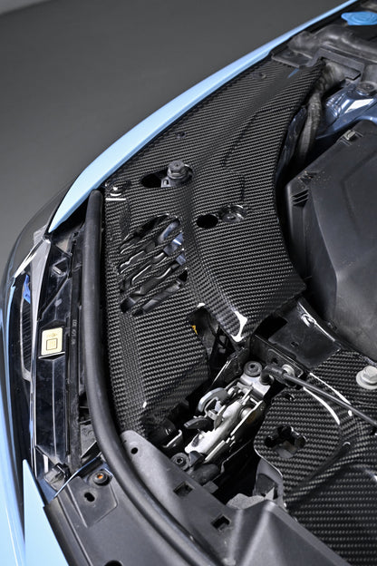 BMW G22 4 Series carbon fiber headlight cover panels are viewed close up along the left edge of the blue engine bay.