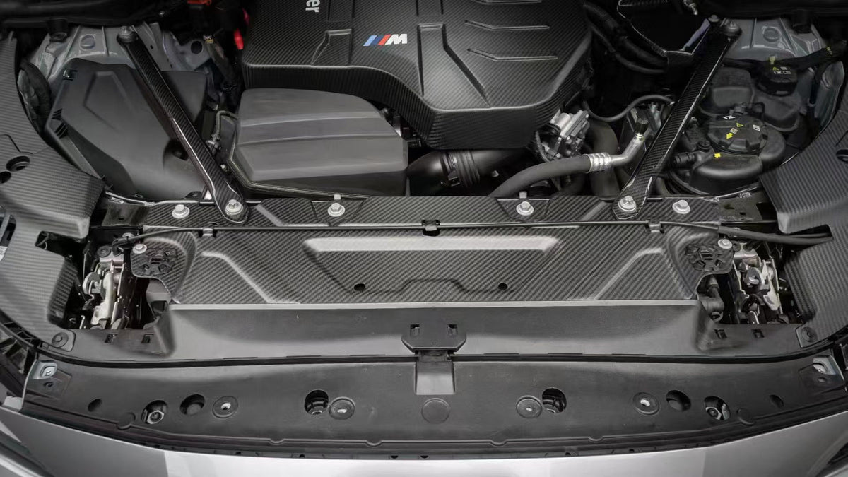 BMW G22 4 Series carbon fiber slam panel cover is seen from the front between the hood latches of a silver car.