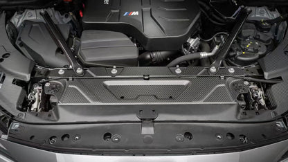 BMW G22 4 Series carbon fiber slam panel cover is seen from the front between the hood latches of a silver car.