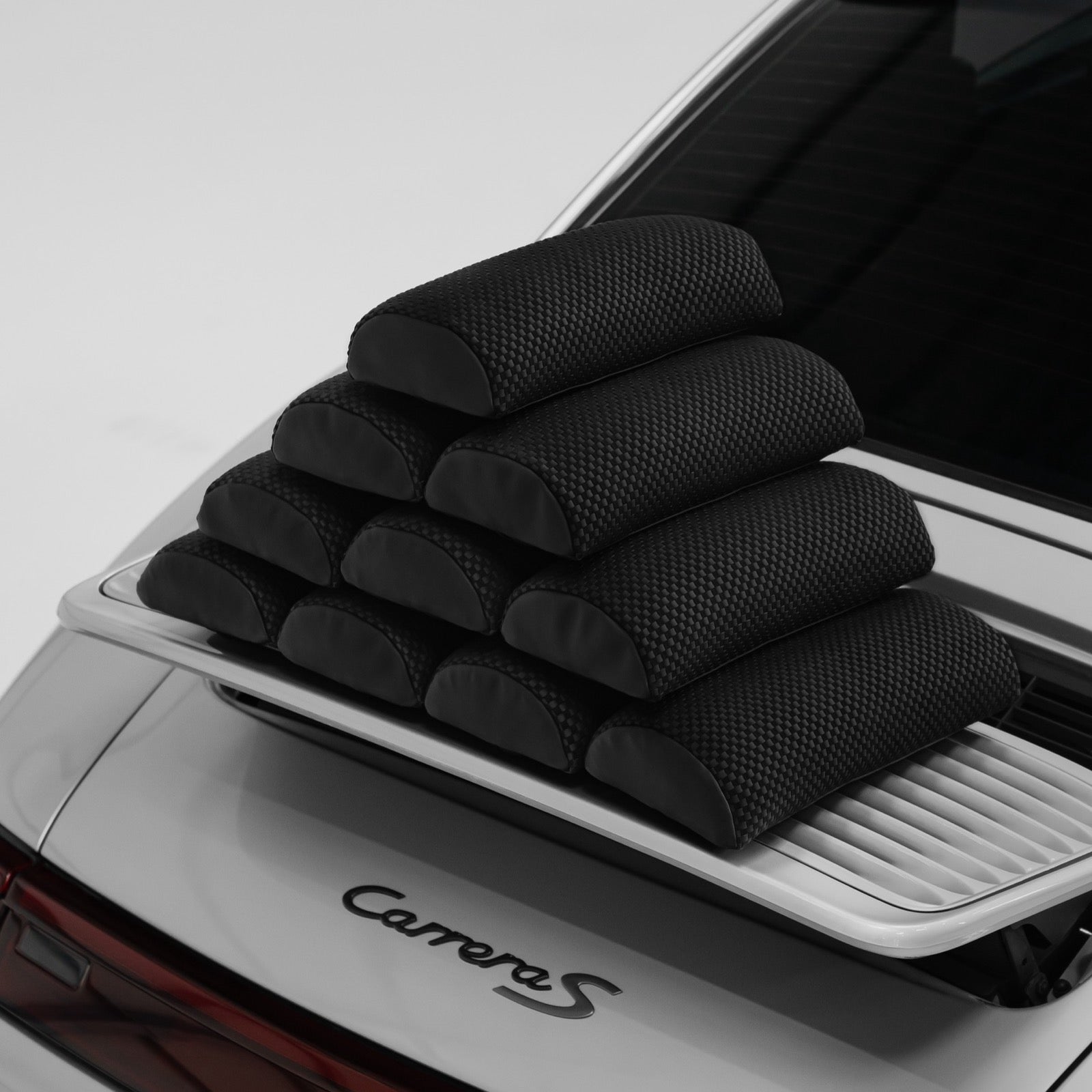 Eight Porsche carbon bucket seat lumbar support pillows stacked on the rear deck of a silver 911 Carrera S