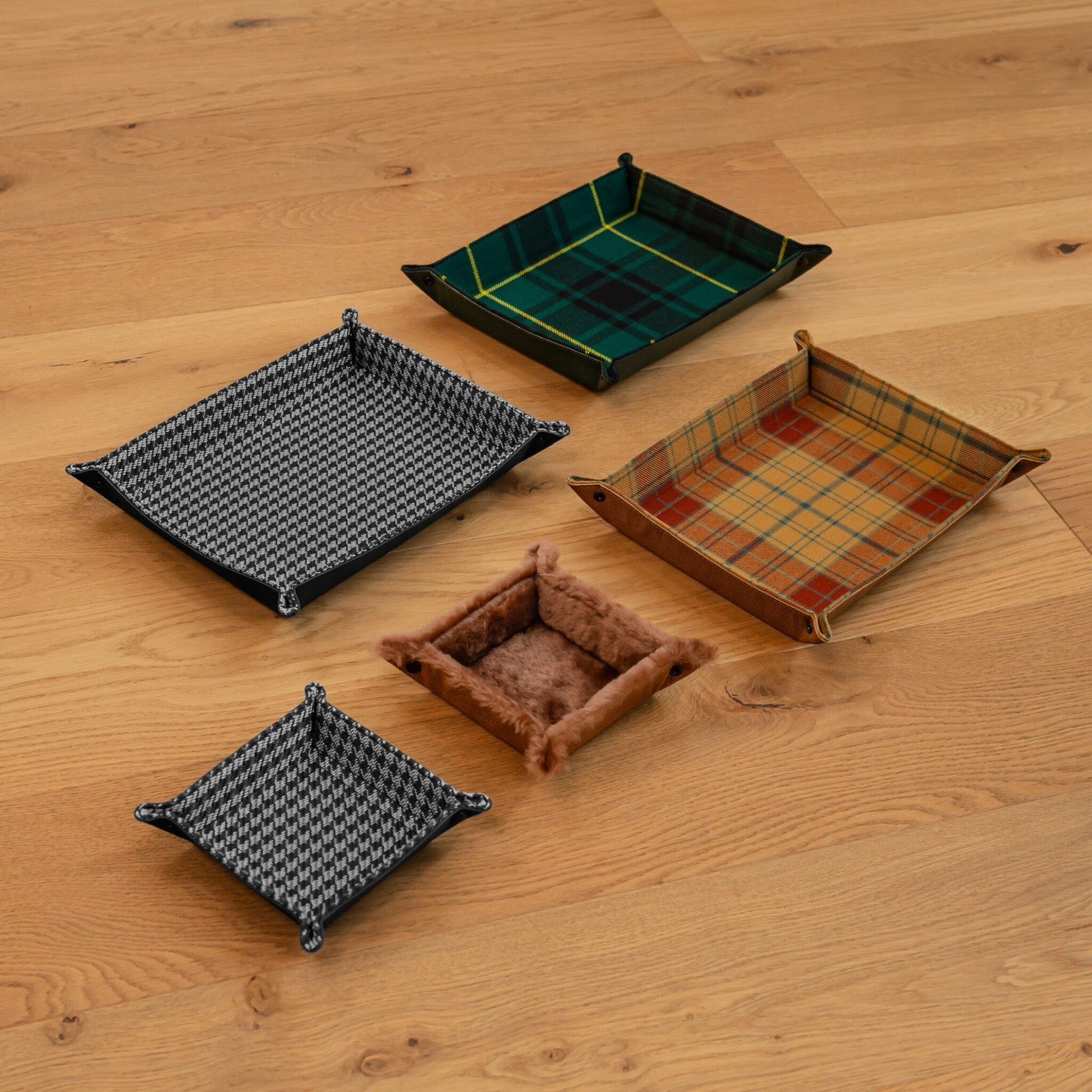 Five handmade leather catch-all trays in tartan, houndstooth and shearling laid out on an oak floor showing small and large sizes